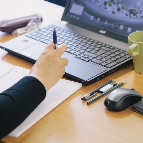 Arm auf einem Notizblock, Hand am Laptop, daneben Tasse, Stifte, Mouse und Handy