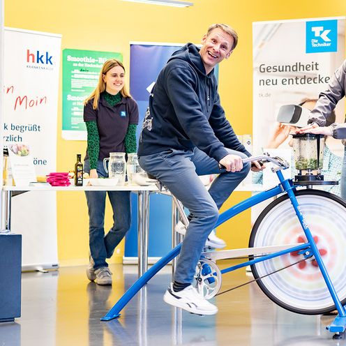 Mann sitzt auf einem Fahrrad, vor ihm ein Smoothiemixer. Rechts und links stehen zwei Frauen. Im Hintergrund stehen Roll-ups und ein Weihnachtsbaum.
