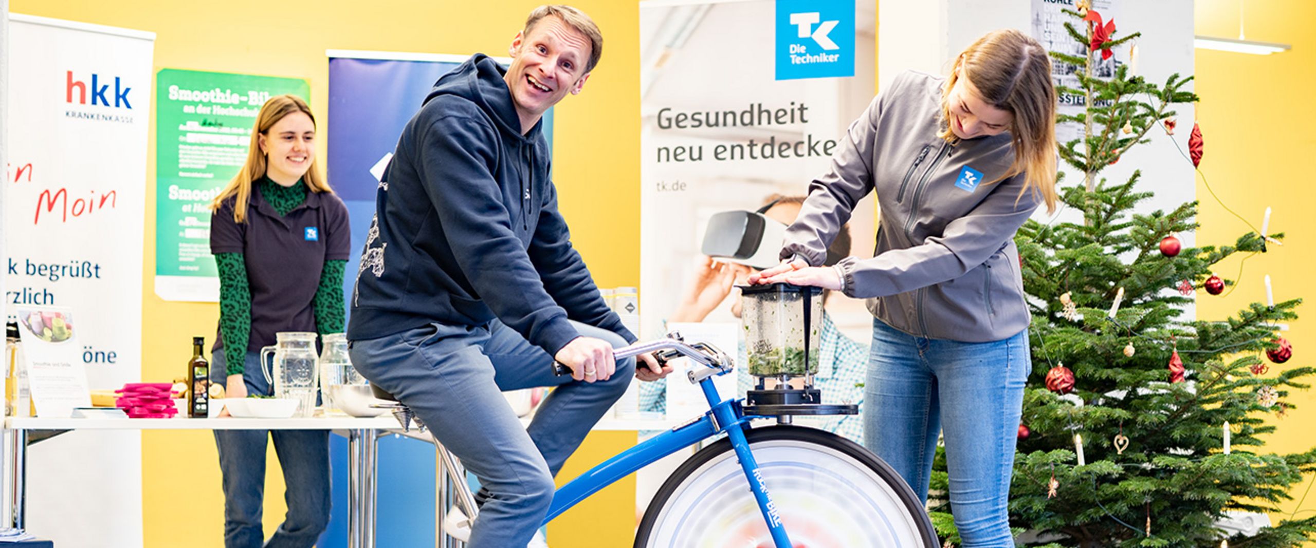 Smoothie-Bike Gesundheitsmanagement Mann sitzt auf einem Fahrrad, vor ihm ein Smoothiemixer. Rechts und links stehen zwei Frauen. Im Hintergrund stehen Roll-ups und ein Weihnachtsbaum.
