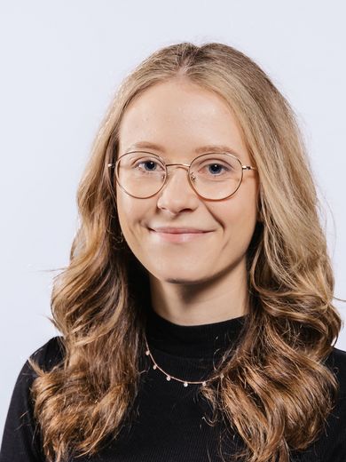 Auf dem Bild ist Kim Stoerterau zu sehen. Sie hat langes blondes Haar, trägt eine runde Brille und einen schwarzen Rollkragenpullover.
