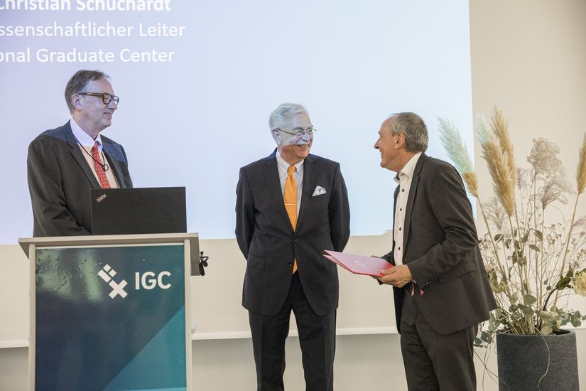 Links steht ein mann am Rednerpult. Rechts &uuml;bergibt ein Mann einem anderen eine Urkunde.