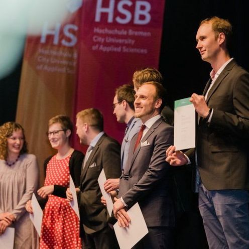 Absolvierende der Hochschule Bremen stehen auf einer B&uuml;hne und halten ihre Abschlussdokumente in den H&auml;nden. 
