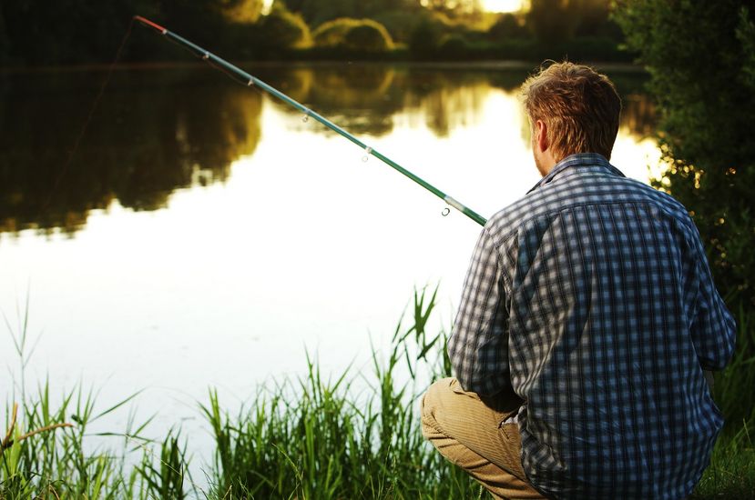 Ein Angler in kariertem Hemd sitzt im Dämmerlicht am Wasser. Der Angler ist von hinten zu sehen.