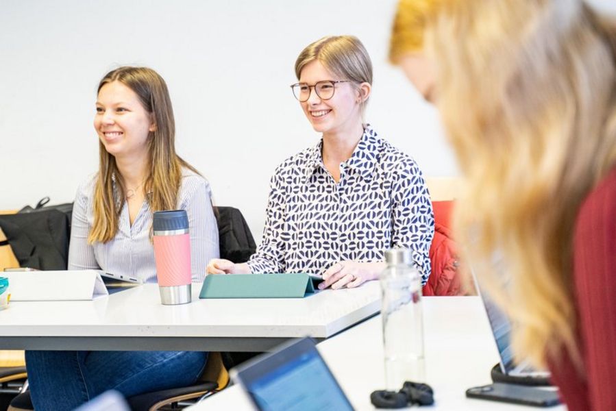 Zwei Studentinnen sitzen nebeneinander an einem Tisch in einem Seminarraum. Sie lachen und schauen nach vorn. Im Vordergrund des Fotos sieht man unscharf eine weitere Studentin von der Seite.