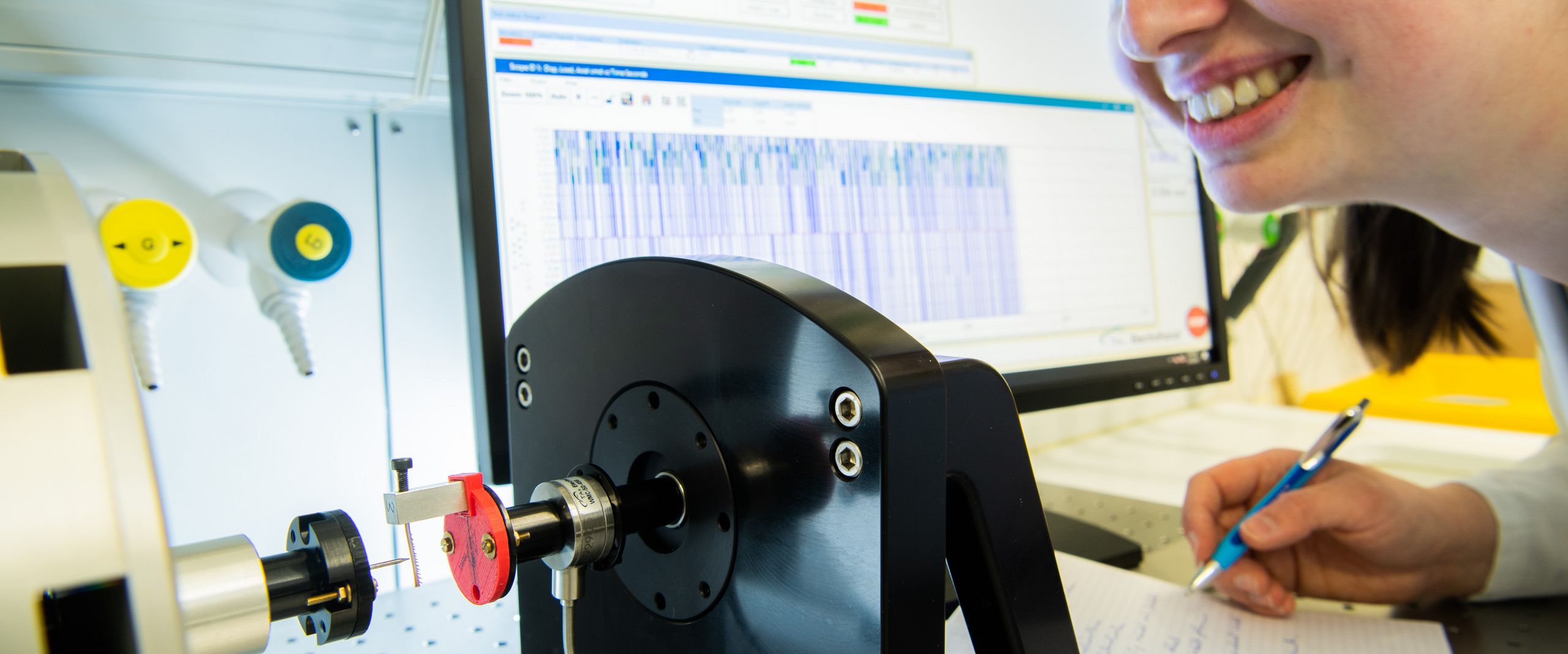 Eine Forscherin beobachtet einen Belastungsversuch mit einem Heuschreckenbein. Im Hintergrund zeigt ein Bildschirm eine Kurve mit den Werten des Versuchs.