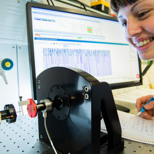 Eine Forscherin beobachtet einen Belastungsversuch mit einem Heuschreckenbein. Im Hintergrund zeigt ein Bildschirm eine Kurve mit den Werten des Versuchs.