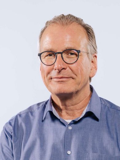 Auf dem Bild ist Andreas Rasch zu sehen. Er hat graues zurück gegeltes Haar und eine runde dunkle Brille. Außerdem trägt er ein blaues Hemd. 