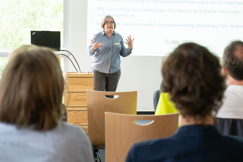 Auf dem Bild ist eine Frau zusehen, die einen Vortrag hält. Im Vordergrund sind unscharf Zuhörende zu erkennen. 