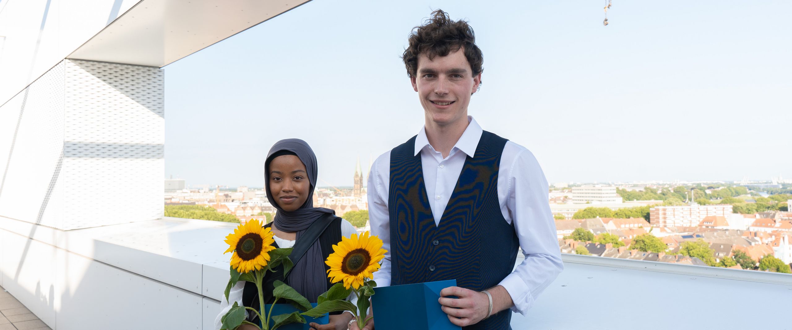 Preisträger:innen der Sommerschau 2024 stehen nebeneiander auf einer Dachterrasse mit Blick auf Bremen. Sie halten Sonnenblumen und eine Urkunde in der Hand und lächeln.