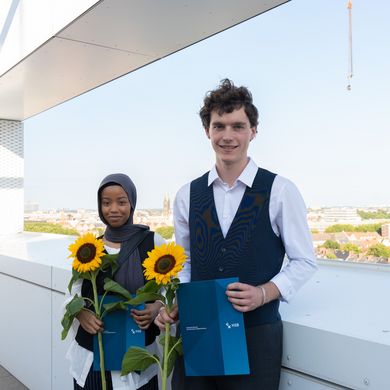 Preisträger:innen der Sommerschau 2024 stehen nebeneiander auf einer Dachterrasse mit Blick auf Bremen. Sie halten Sonnenblumen und eine Urkunde in der Hand und lächeln.