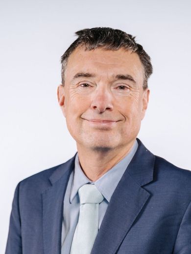 Auf dem Bild ist Jens Eschenbächer zu sehen. Er hat kurzes dunkles Haar und trägt ein hellblaues Hemd mit leicht grünlicher Krawatte und einem dunkelblauen Jacket. 