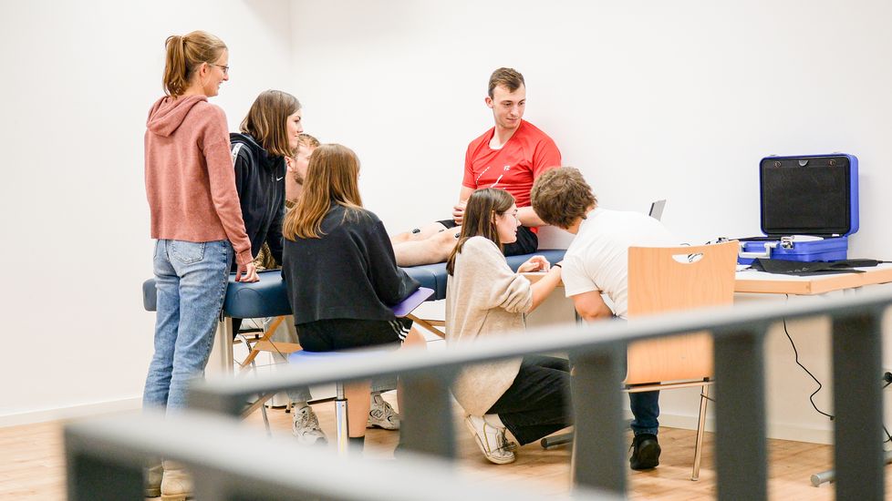Mehrere Studierende sitzen um eine Behandlungsliege. Ein Student hat Elektroden auf dem Bein und das elektrische Signal wir auf einem Monitor angezeigt.