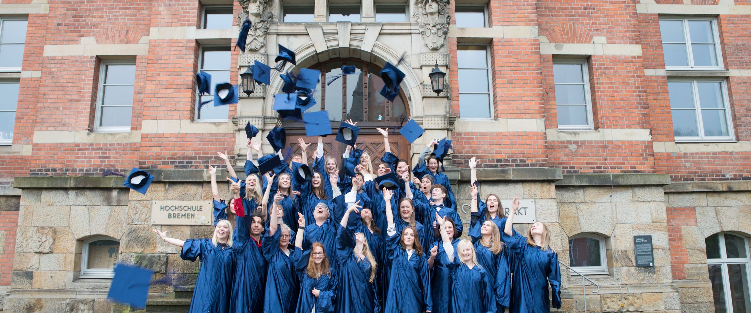 Absolventinnen und Absolventen des Studiengangs stehen auf den Treppen vor dem Eingangsportal des alten Hochschulgebäudes in Talaren und werfen lachend die Hüte als Zeichen ihrer erfolgreichen Graduation in die Luft.