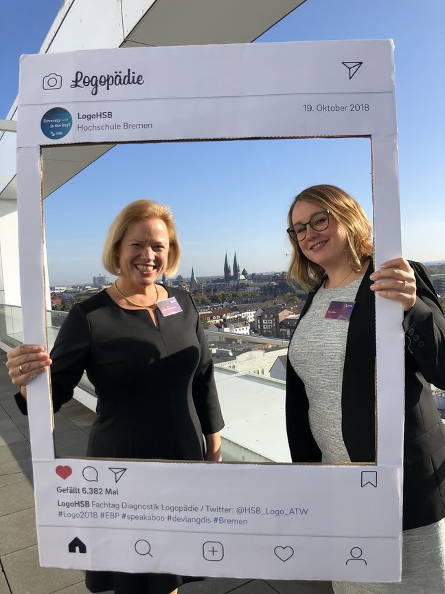 Wir vermitteln Logopädie Professorin und Mitarbeiterin stehen auf dem Dach der Hochschule Bremen und halten einen Instagram-Post als rahmend für die Aufnahme zum Fotografen. Im Hintergrund sieht man die Bremer Innenstadt gekennzeichnet durch die Türme des Bremer Doms.